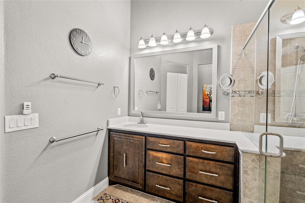 Bathroom featuring wood-finish cabinetry, a neutral stone-surface countertop, a large framed mirror with six-bulb vanity lighting, and a glass-enclosed shower with tiled surround
