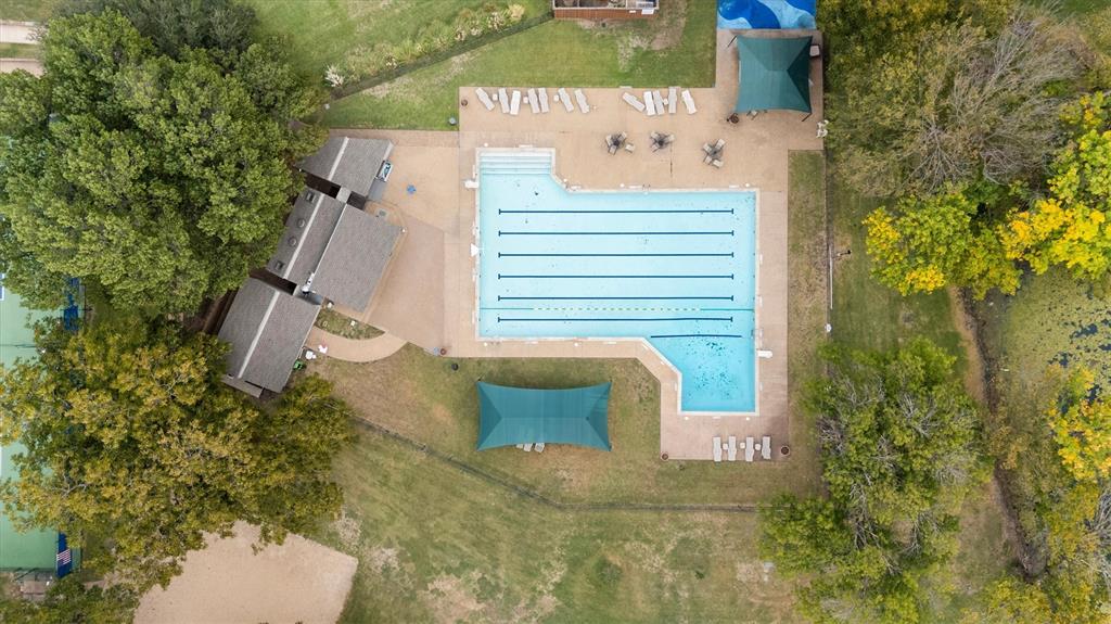 Aerial view of a swimming pool with multiple lap lanes, paved pool deck, shaded canopy structures, and mature tree-lined perimeter