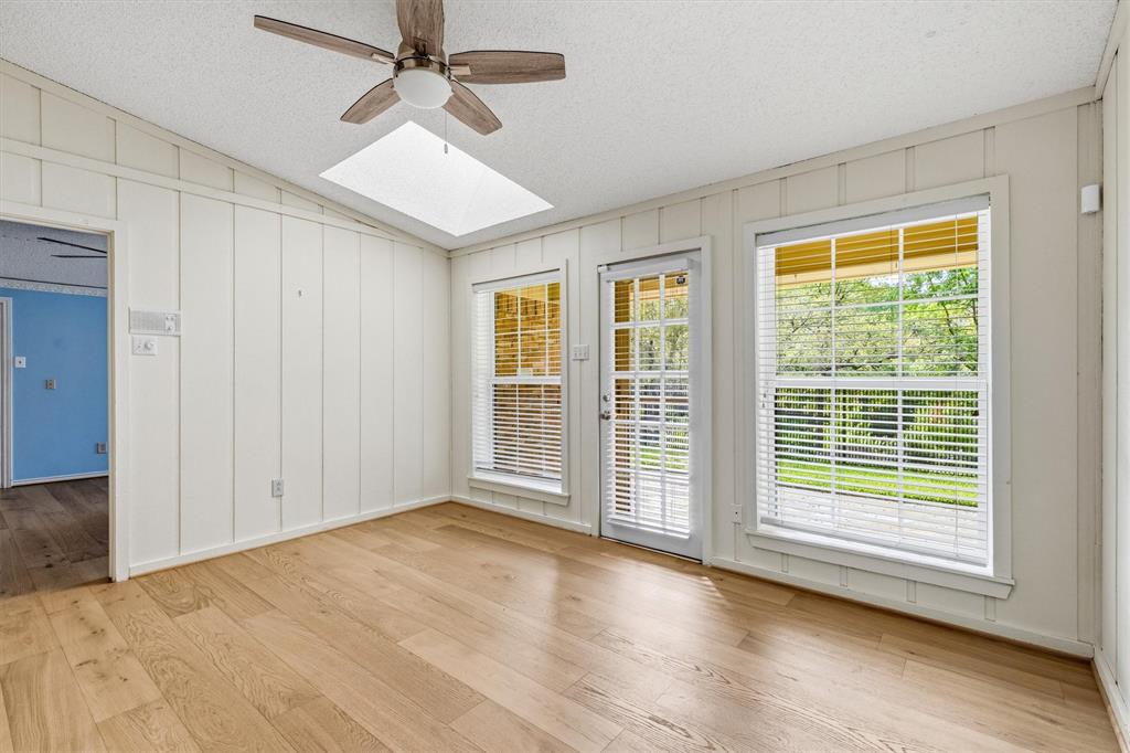 Vaulted room featuring wood-finish flooring, vertical wall paneling, a ceiling fan, and a skylight, with a glass-paneled exterior door flanked by two windows
