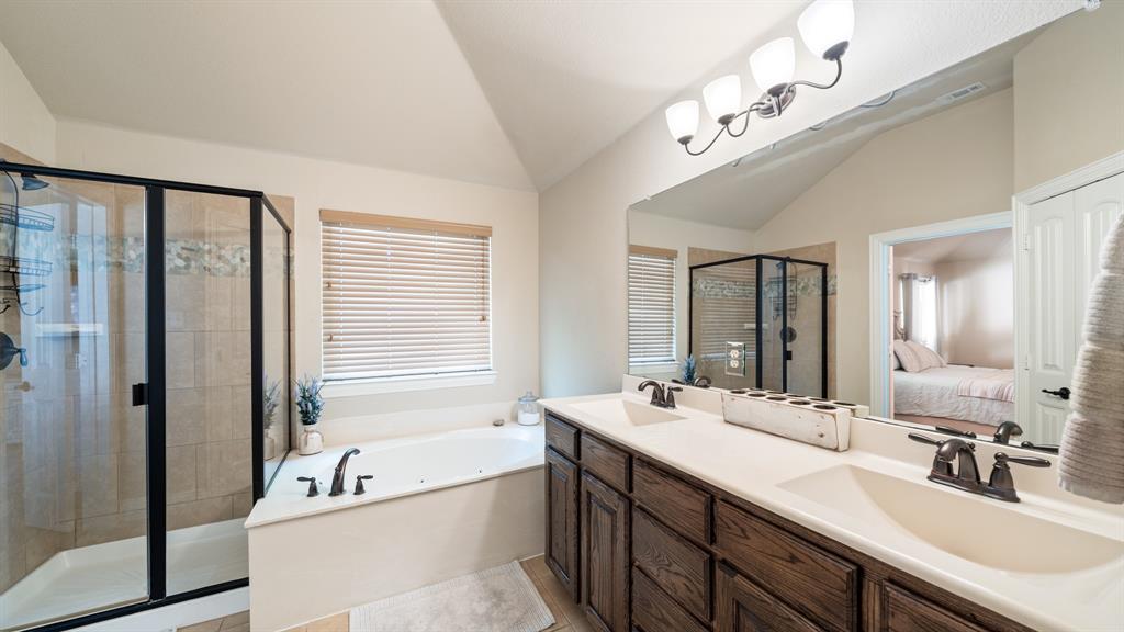 Bathroom featuring a dual-sink vanity with dark-finish cabinetry, a large wall-mounted mirror, a jetted soaking tub, and a separate glass-enclosed shower with tiled surround
