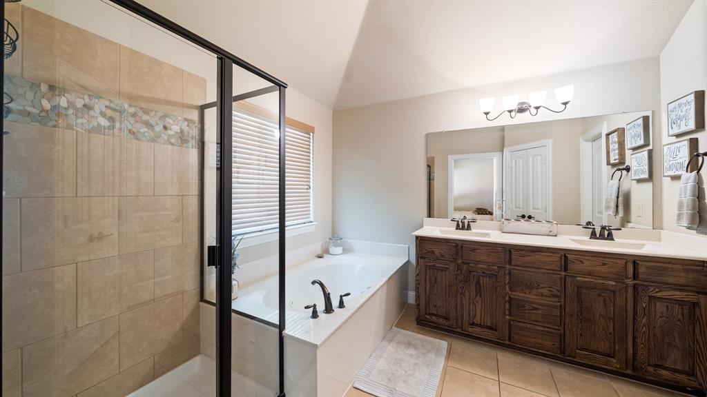 Dual vanity with dark wood-finish cabinetry, soaking tub, glass-enclosed shower with tiled walls and decorative stone inlay, and wall-mounted light fixture