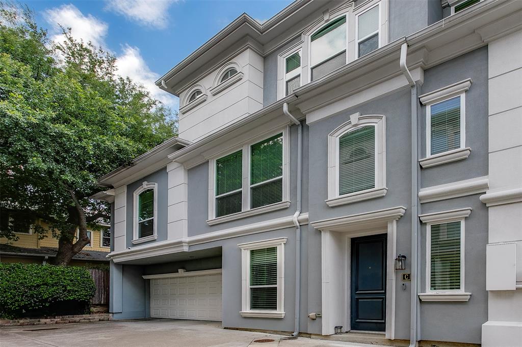 Stucco exterior featuring multiple windows with decorative trim, a dark paneled front door, an integrated garage with a white door, and mature trees providing natural shading