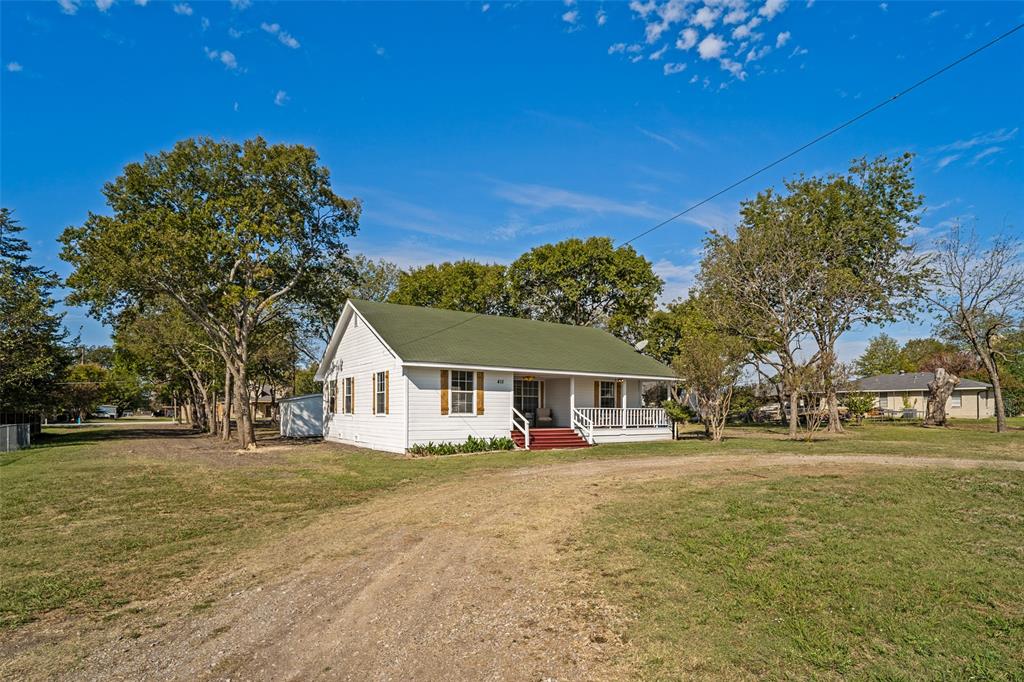 Exterior featuring white siding, a green gabled roof, an expansive lawn, mature trees, and a covered front porch with a wood-finish staircase