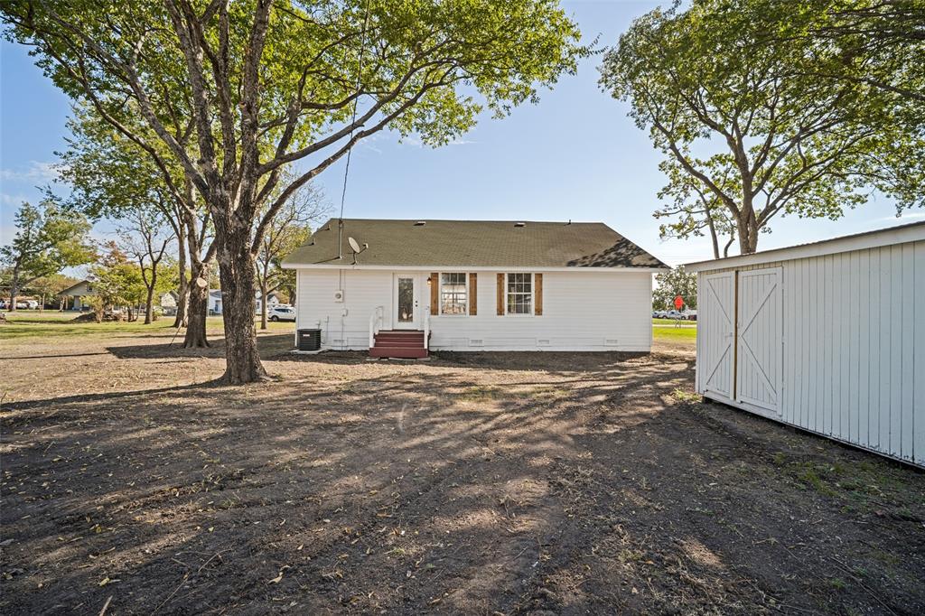 White exterior siding, shingle roofing, wooden window shutters, and a detached storage structure on a spacious lot with mature trees