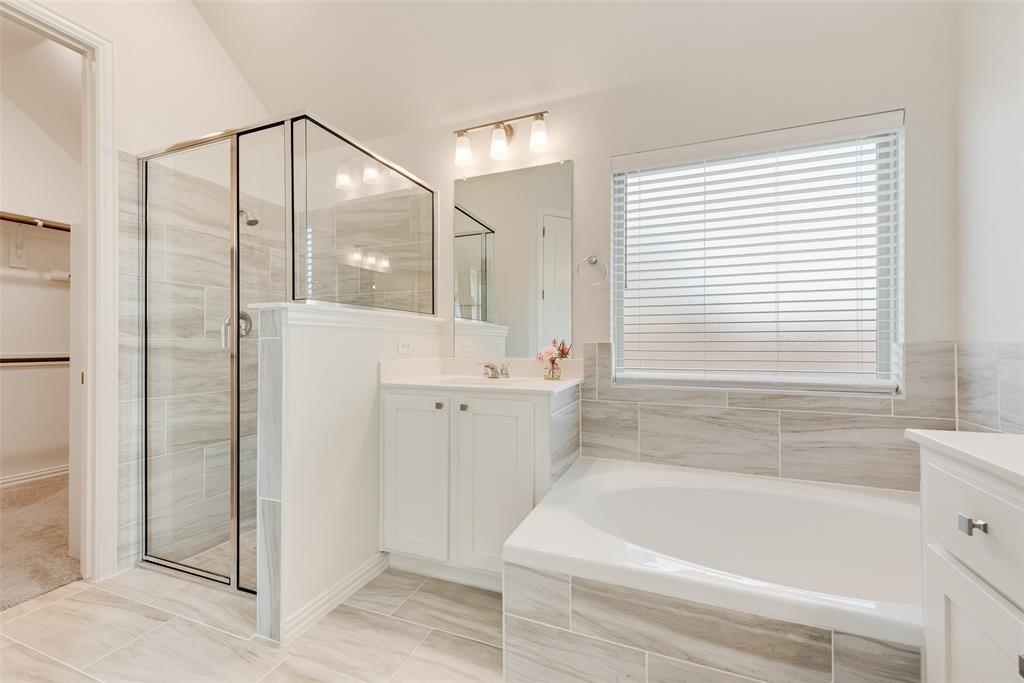 Bathroom featuring a walk-in glass-enclosed shower, soaking tub with tile surround, white cabinetry, rectangular mirror, and light-tone tile flooring
