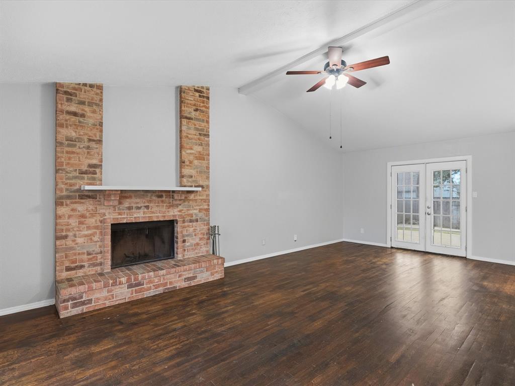 Vaulted ceiling living area featuring a full-height brick fireplace, wood-finish flooring, a ceiling fan with integrated lighting, and glass-paned double doors