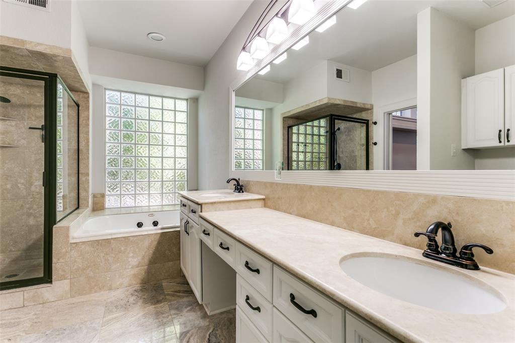 Dual vanity featuring white cabinetry, stone-surface countertops, and dark hardware, complemented by a glass-block window, deep soaking tub, and a tiled walk-in shower