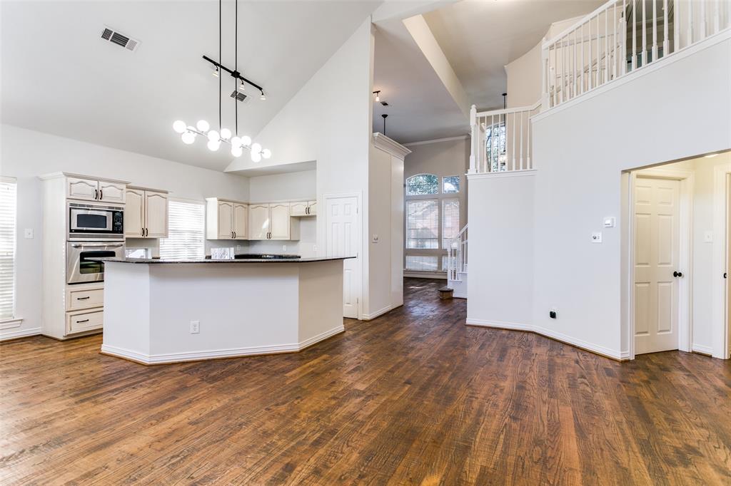 Open-concept interior featuring vaulted ceilings, wood-finish flooring, a kitchen island with stone countertops, built-in stainless steel appliances, and an elevated wooden staircase with white balusters