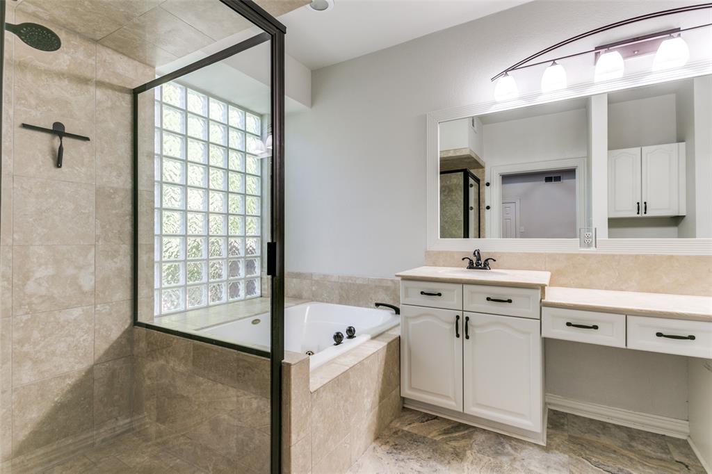 Bathroom featuring a jetted soaking tub, glass-enclosed shower with tile surround, white cabinetry, neutral stone-tile flooring, and a glass block window
