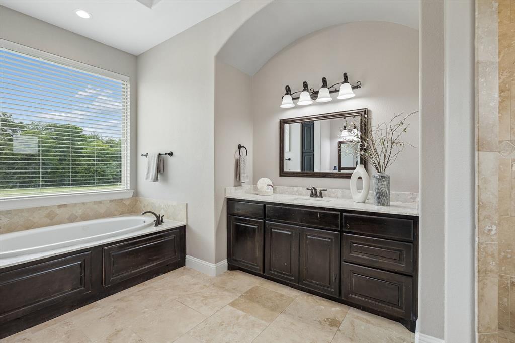 Bathroom featuring a recessed tub with stone tile surround, stone-topped vanity with dark wood cabinetry, and a large wall-mounted mirror with four-light fixture