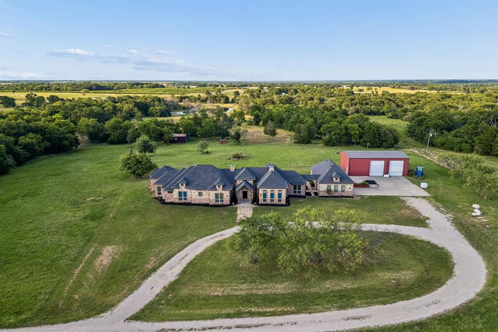 Aerial perspective of a multi-gabled stone and brick exterior, expansive acreage with mature trees, detached metal workshop with concrete driveway, and a gravel circular motor court