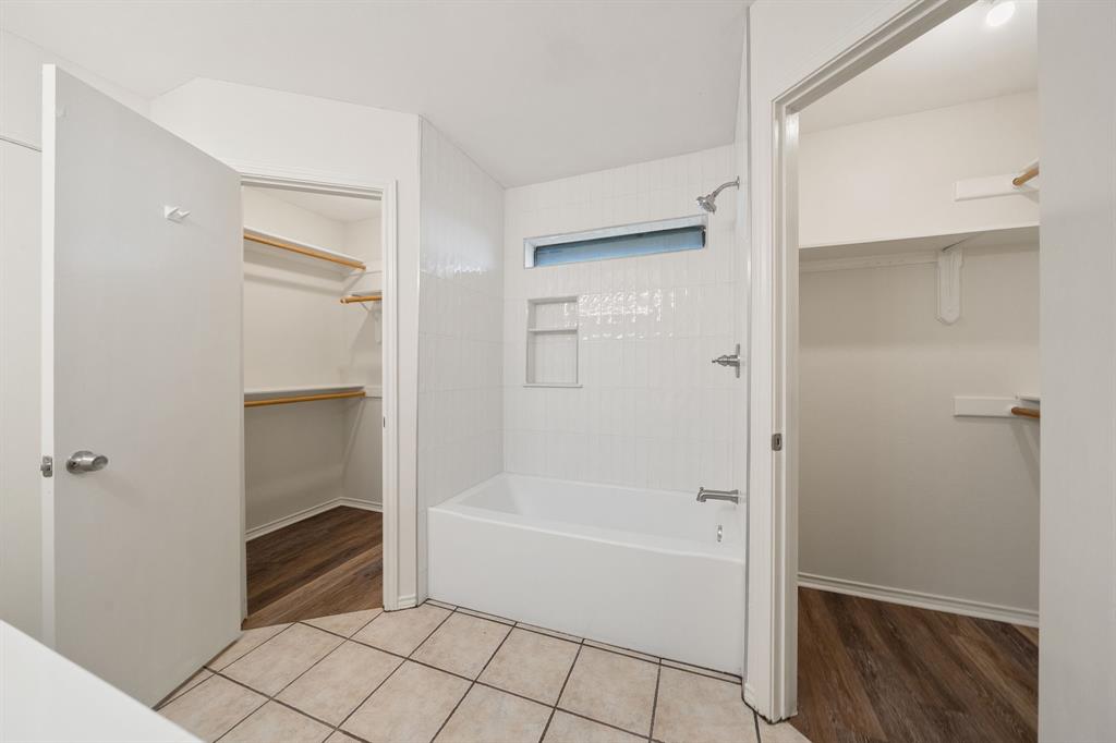 Bathroom featuring a white tile tub surround with built-in recessed shelving, clerestory window, metal shower fixture, ceramic tile flooring, and dual walk-in closets with wood-finish flooring