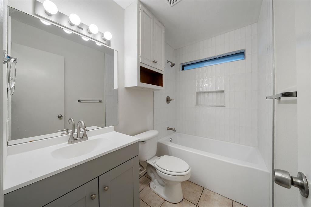 Bathroom featuring a grey vanity with a solid-surface countertop, white tile tub surround with a built-in niche, clerestory window, and over-mirror light fixture
