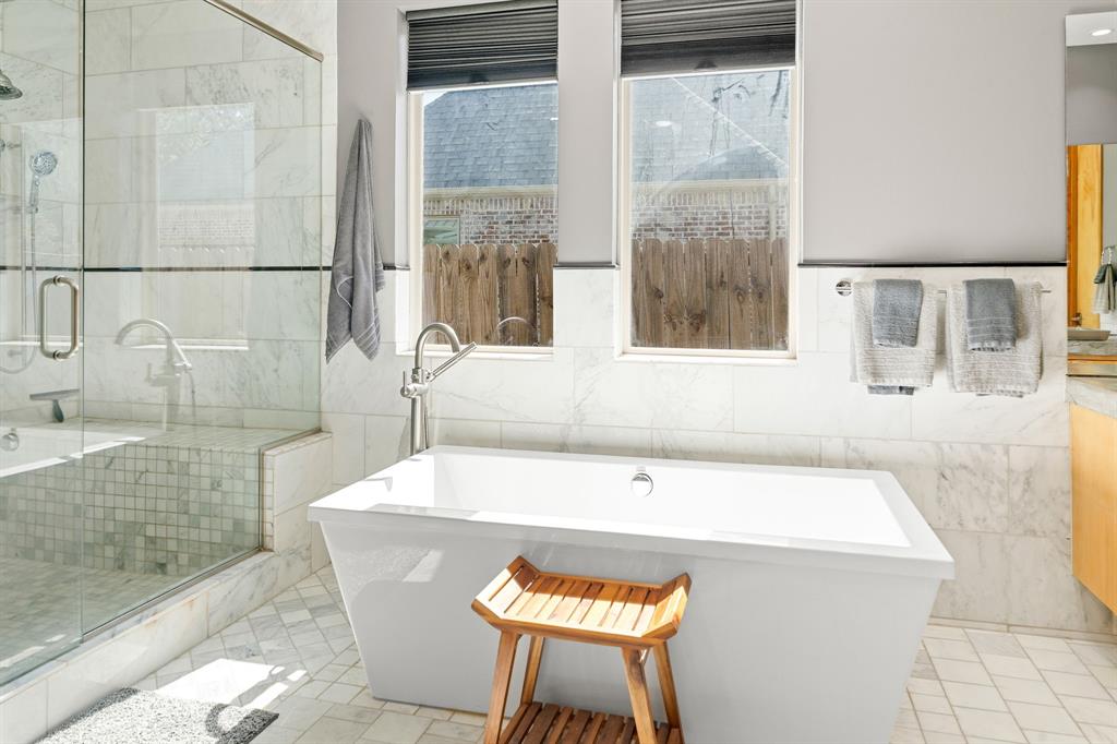 Freestanding soaking tub with chrome fixtures, glass-enclosed shower, stone wall tile, and light-toned floor tile