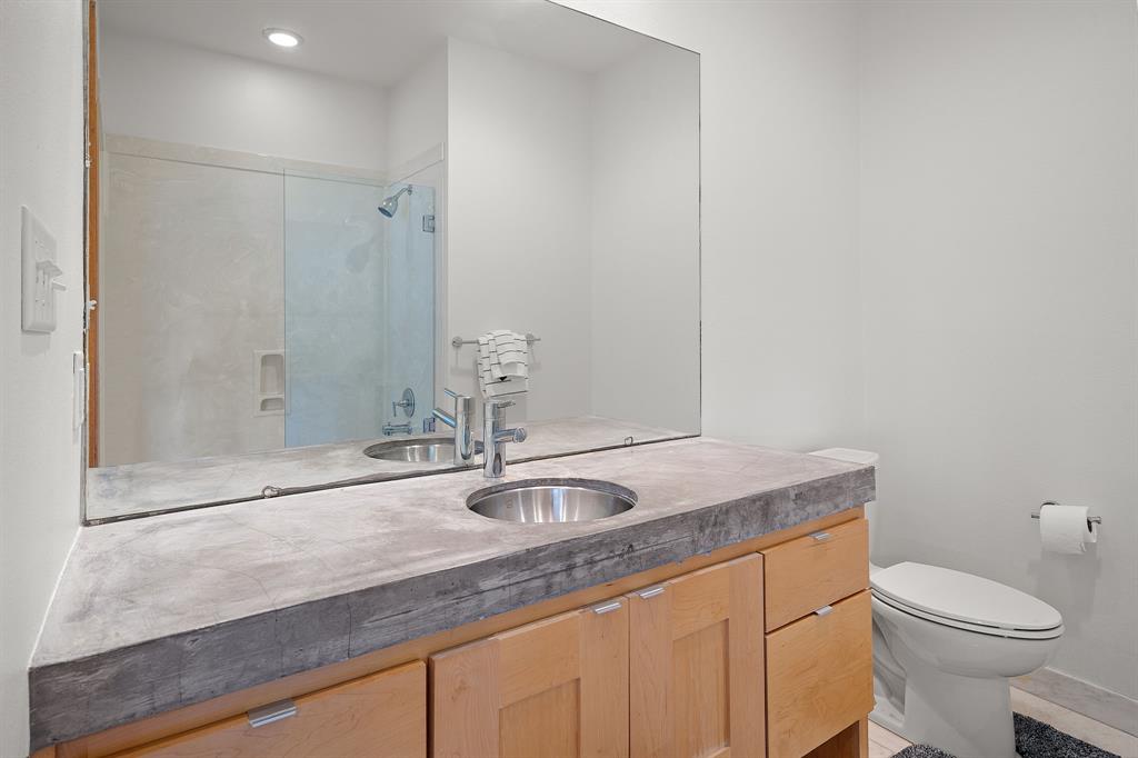 Bathroom featuring a concrete-style countertop, light wood cabinetry, a large wall-mounted mirror, an undermount circular sink with a chrome faucet, and a shower with a glass enclosure