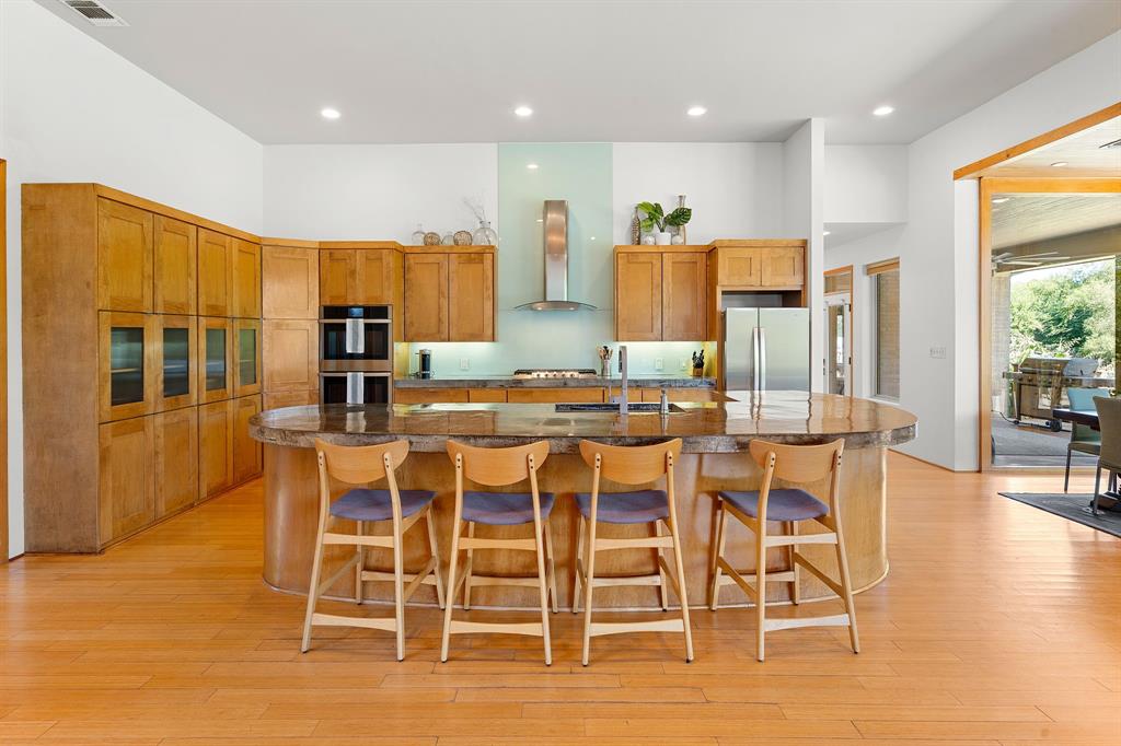 Kitchen featuring wood cabinetry, curved stone-topped island with seating, stainless steel double wall ovens, stainless steel range hood, and wood-finish flooring