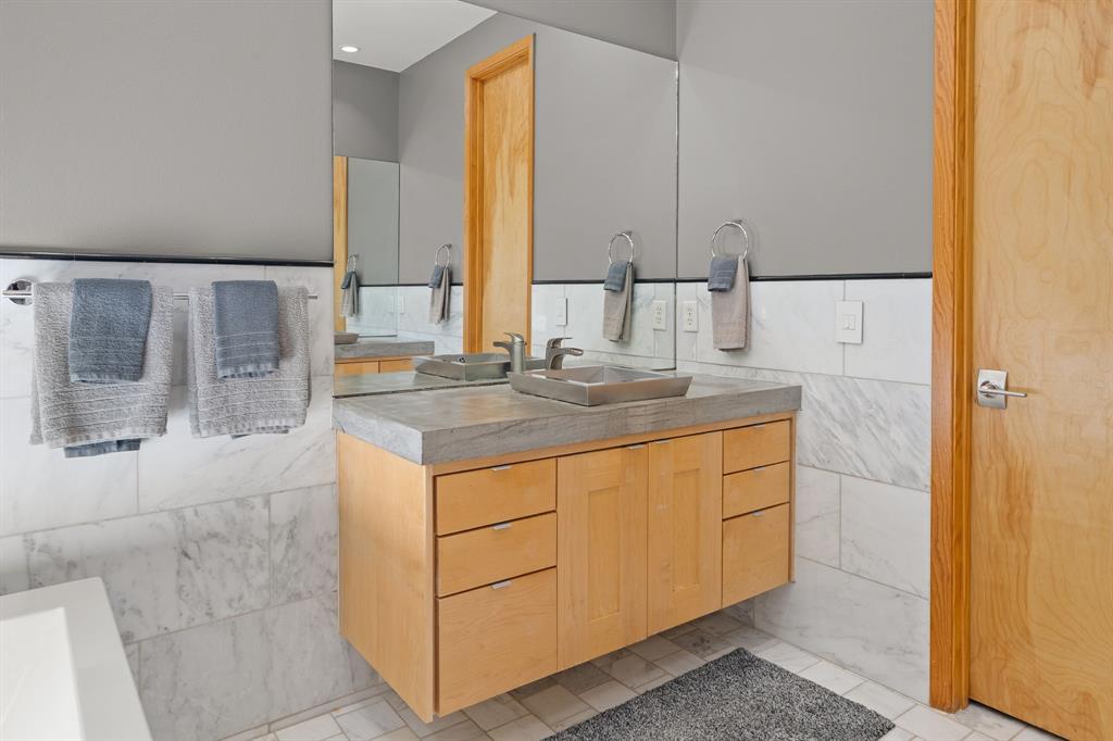 Floating light wood vanity featuring a concrete-style countertop, integrated vessel sink, polished metal hardware, and large-format wall tiling