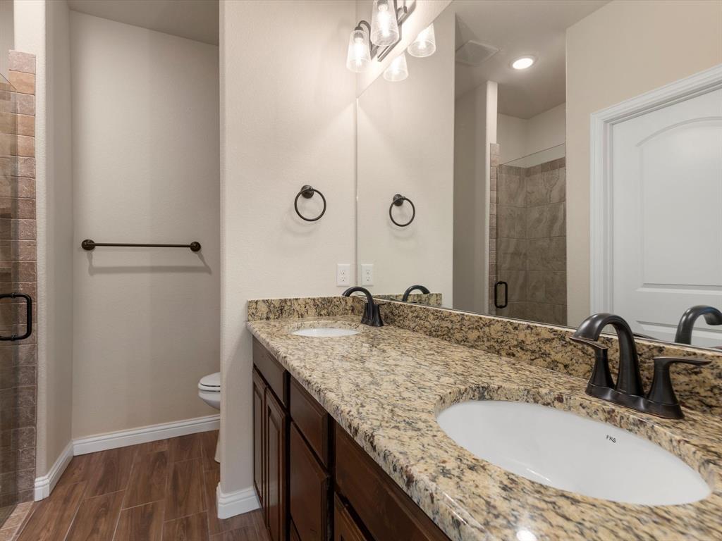 Double-vanity bathroom featuring stone-patterned countertops, dark wood-finish cabinetry, wood-finish flooring, oil-rubbed bronze fixtures, and a glass-enclosed shower