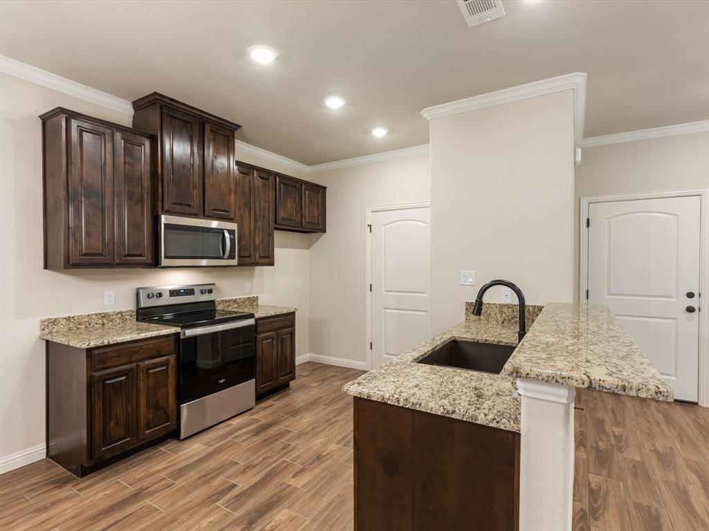 Kitchen featuring stained wood cabinetry, stone countertops, stainless steel appliances, a center island with a drop-in sink, and wood-finish flooring Kitchen featuring stained wood cabinetry, stone countertops, stainless steel appliances, a center island with a drop-in sink, and wood-finish flooring