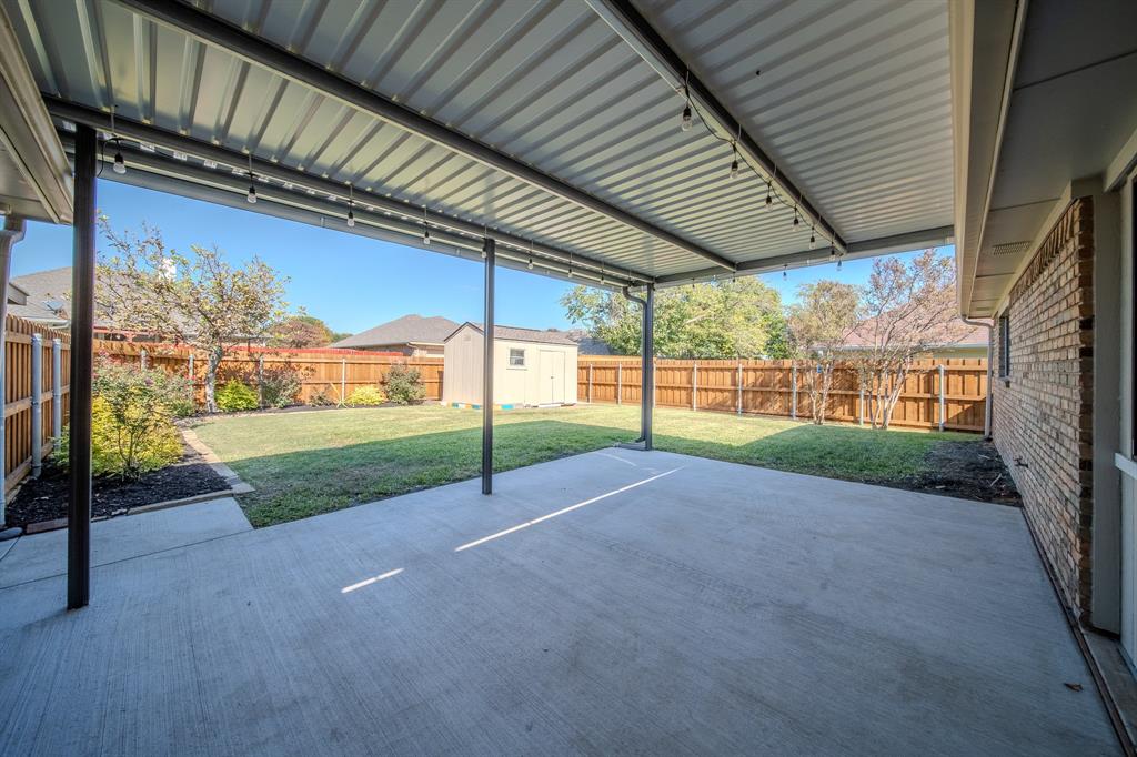 Covered concrete patio with a metal roof, wood-plank privacy fencing, and a detached storage structure set within a grass lawn