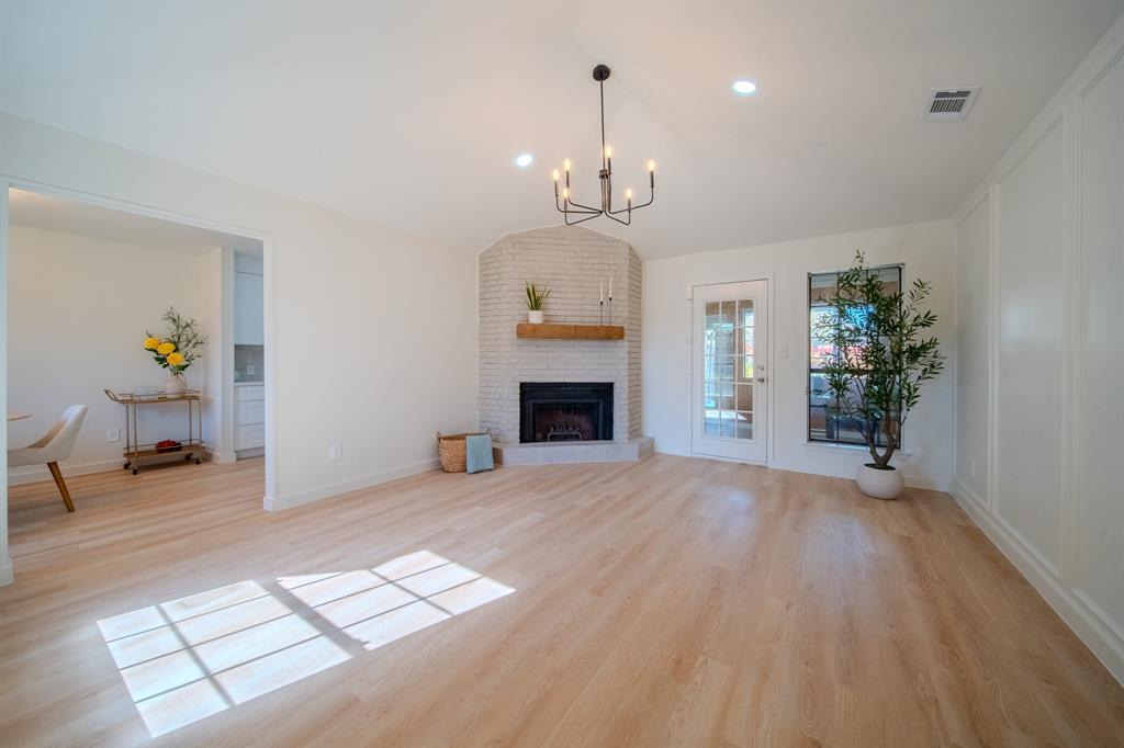 Open living area featuring light-toned wood-finish flooring, a corner brick fireplace with a natural wood mantel, recessed lighting, a modern multi-arm chandelier, and decorative wall molding