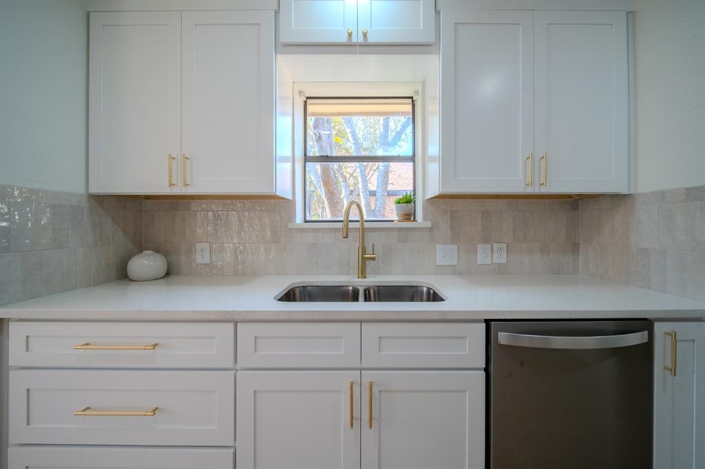 Shaker-style cabinetry with brass hardware, solid surface countertops, metallic finish faucet, stainless steel dishwasher, and textured tile backsplash