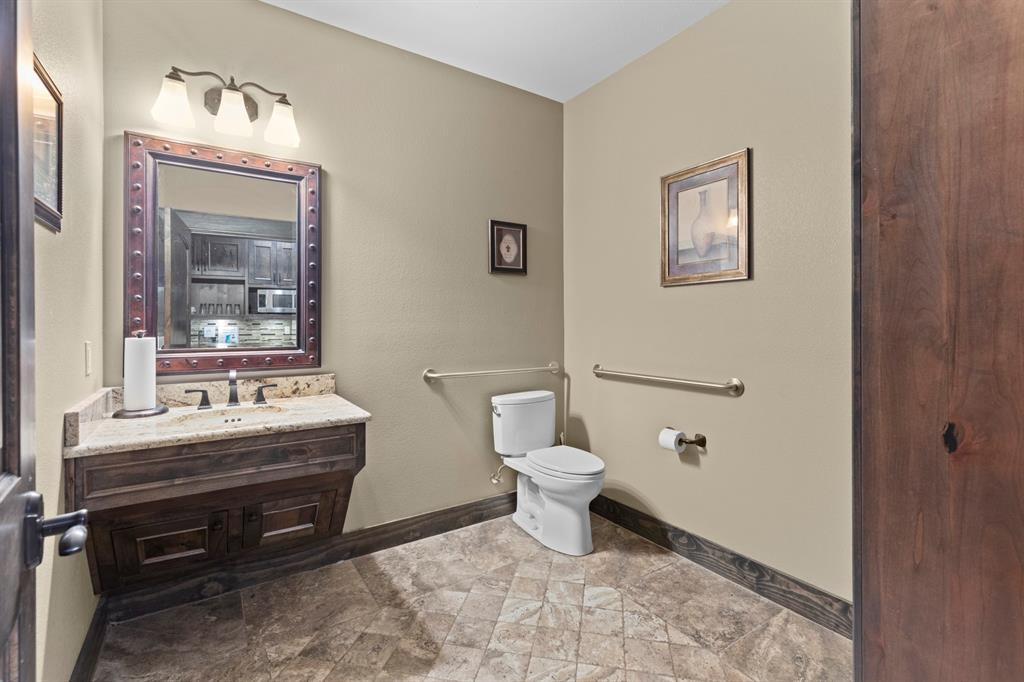 Custom wooden vanity with stone countertop, decorative metal-trimmed mirror, three-light fixture, and stone-tile flooring