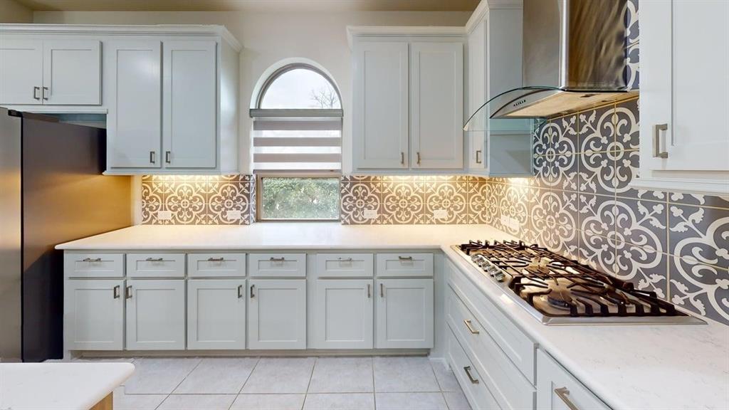 Kitchen featuring white shaker-style cabinetry, patterned tile backsplash, solid-surface countertops, gas cooktop with stainless steel range hood, and arched window
