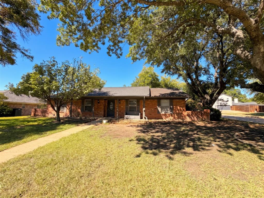 Single-story brick exterior featuring a low-pitch roof, decorative brick privacy wall, and mature shade trees