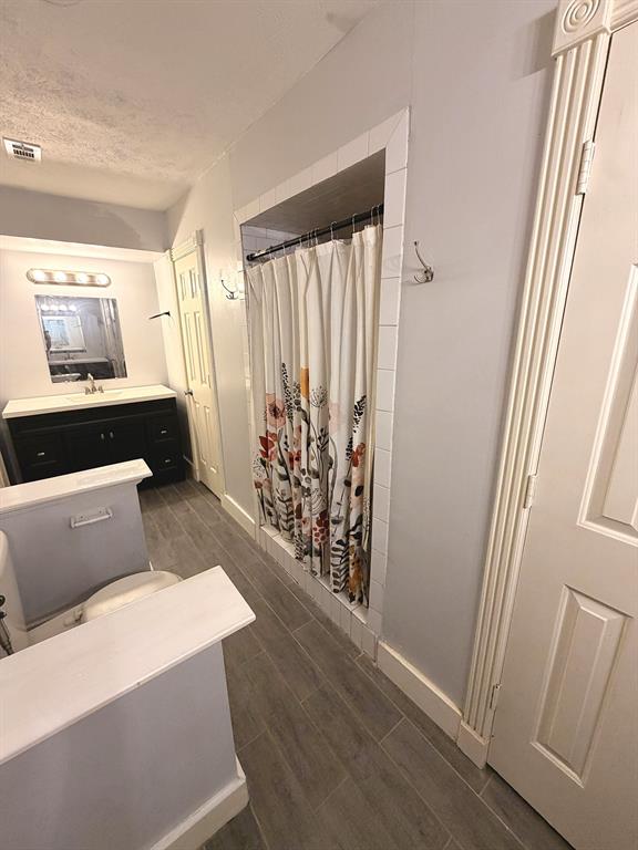 Bathroom featuring wood-finish tile flooring, a dark-toned vanity with a white countertop, a recessed shower area with white tile surround, and two panel-style doors