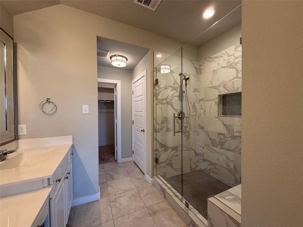 Walk-in shower featuring large-format tile surround, glass enclosure, built-in recessed niche, and stone-finish floor tiling, complemented by a white vanity with integrated basin