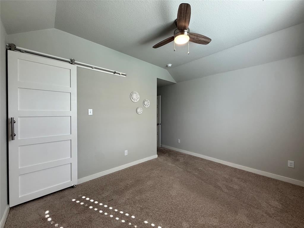 Vaulted ceilings, carpeted flooring, and a white sliding barn door featuring a stainless steel track and hardware, complemented by a ceiling fan with integrated lighting Vaulted ceilings, carpeted flooring, and a white sliding barn door featuring a stainless steel track and hardware, complemented by a ceiling fan with integrated lighting