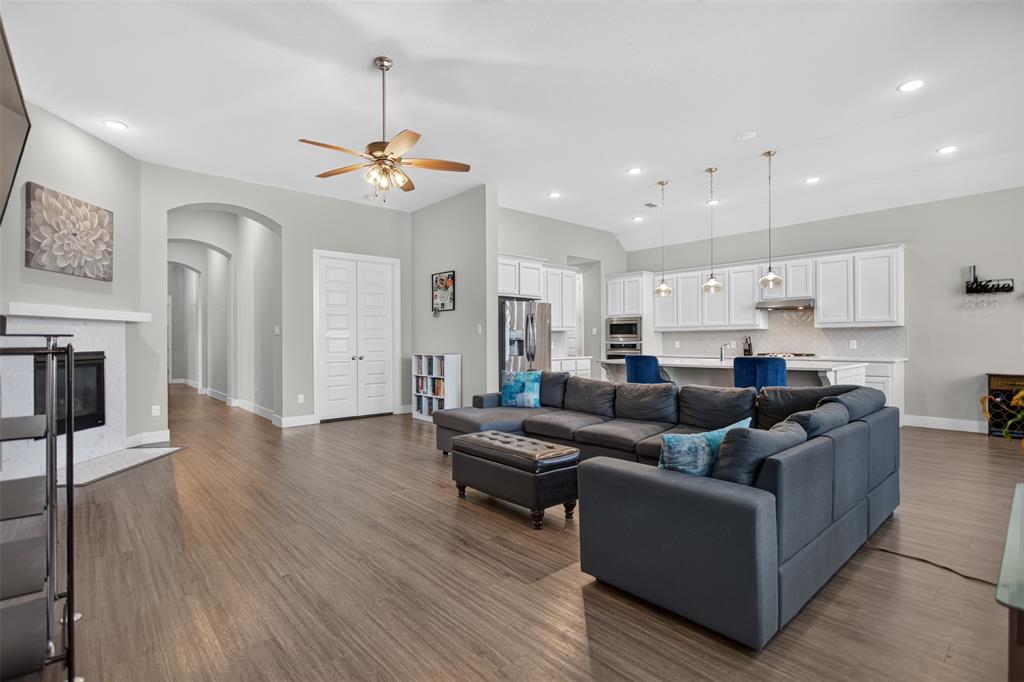 Open-concept living space featuring wood-finish flooring, high ceilings with recessed lighting, a stone-surround fireplace, and a kitchen equipped with white cabinetry and a central island with pendant lighting