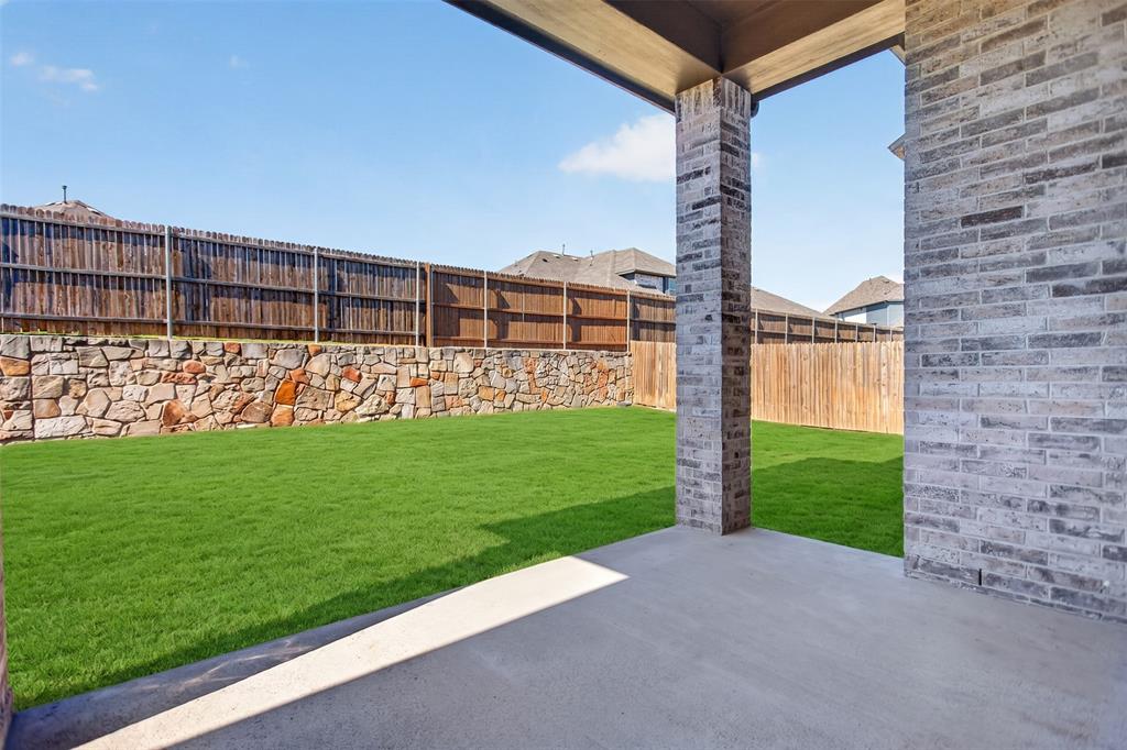 Covered patio featuring brick pillar supports, concrete flooring, manicured lawn, stone retaining wall, and wood fencing