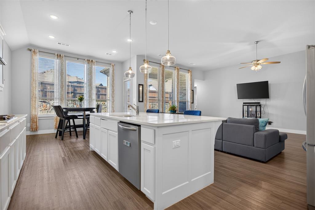 Kitchen featuring a large central island with white cabinetry, light-toned stone-finish countertop, stainless steel dishwasher, and three glass pendant lights, complemented by wood-finish flooring and multiple tall windows