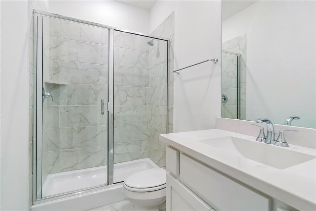 Bathroom featuring a walk-in shower with glass-enclosure doors, light-toned stone-finish tile surround, built-in shelving, white cabinetry with a solid-surface countertop, and a wall-mounted mirror
