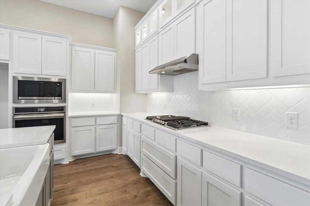 Kitchen featuring white shaker-style cabinetry with glass-front uppers, solid surface countertops, a herringbone tile backsplash, integrated stainless steel appliances, and wood-finish flooring