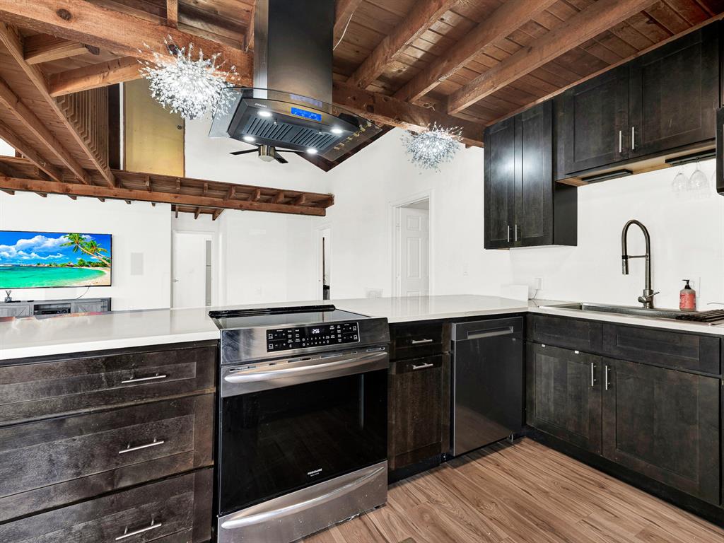 Kitchen featuring dark wood cabinetry, solid surface countertops, stainless steel appliances, a center-mounted range hood, and an exposed beam vaulted ceiling