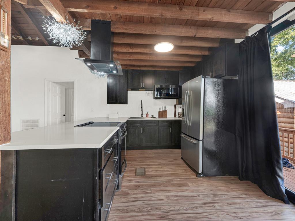 Kitchen featuring dark cabinetry, quartz countertops, a central island with a built-in cooktop, a stainless steel refrigerator, and an exposed wood-beam ceiling