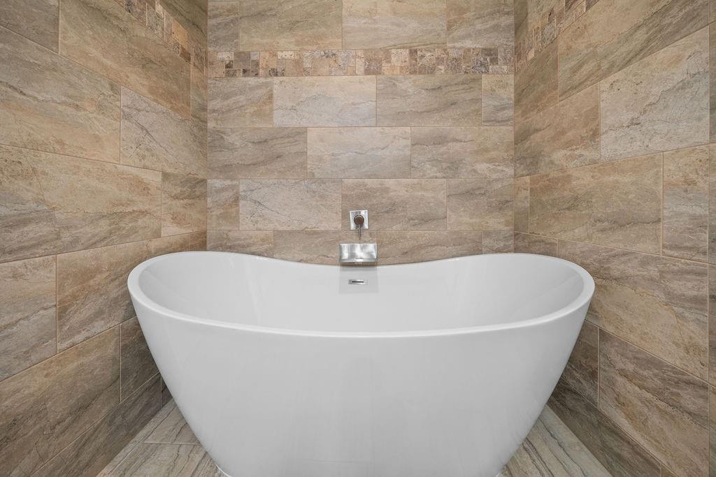 Freestanding white soaking tub featuring a wall-mounted faucet and surround of large-format stone-finish tile with integrated mosaic accent banding