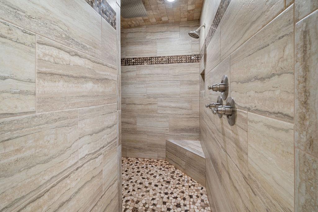 Walk-in shower featuring large-format neutral-toned wall tile, decorative mosaic tile accents, a built-in corner bench, a ceiling-mounted rainfall showerhead, and a multi-handle faucet system
