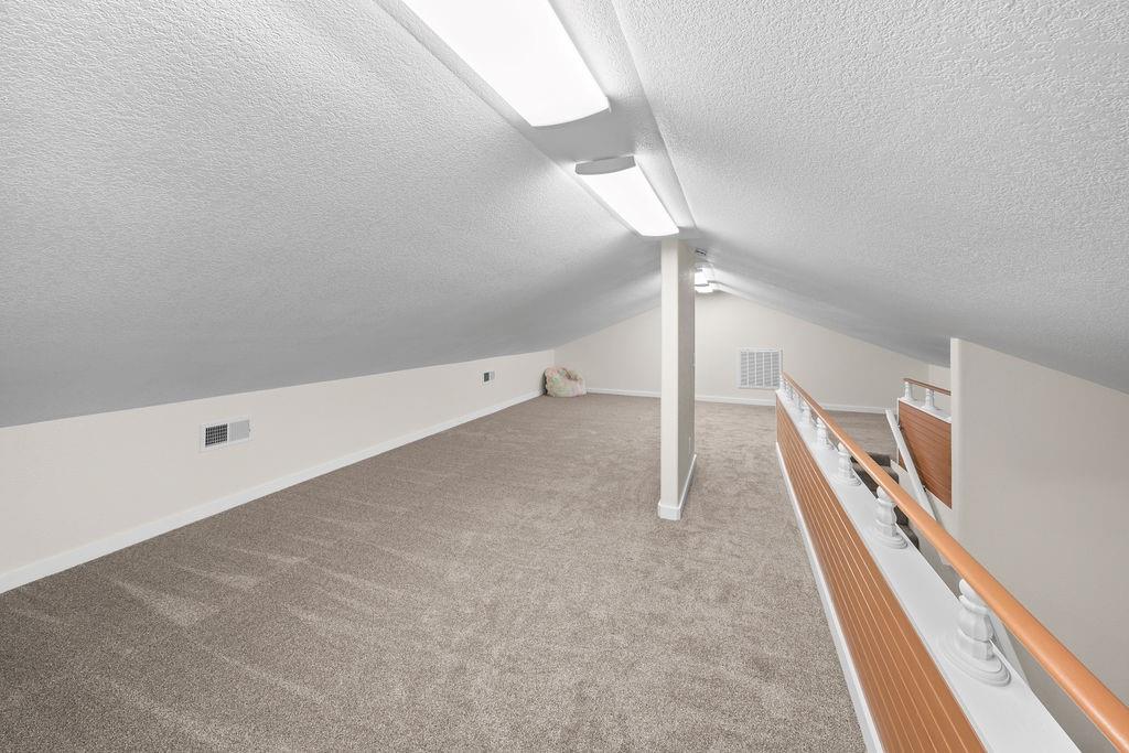 Finished loft space featuring angled ceilings, wall-to-wall carpeting, flush-mount lighting, and a white balustrade with a wooden handrail