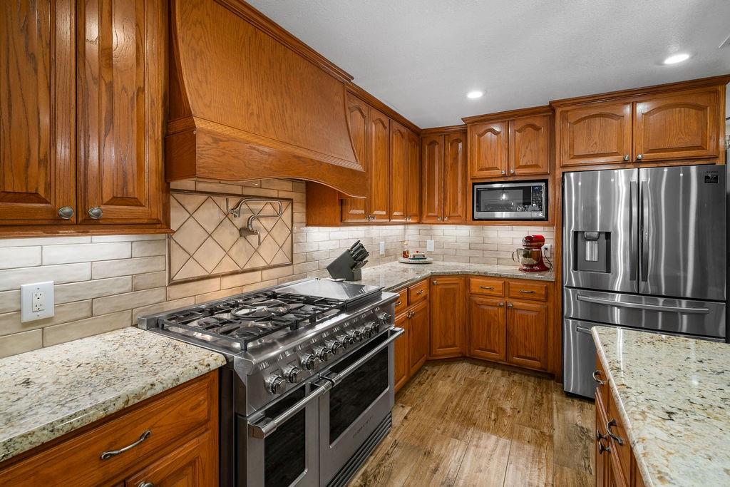 Kitchen featuring raised-panel wood cabinetry, natural stone countertops, a multi-burner stainless steel gas range with a custom wood-enclosed vent hood, and wood-finish flooring