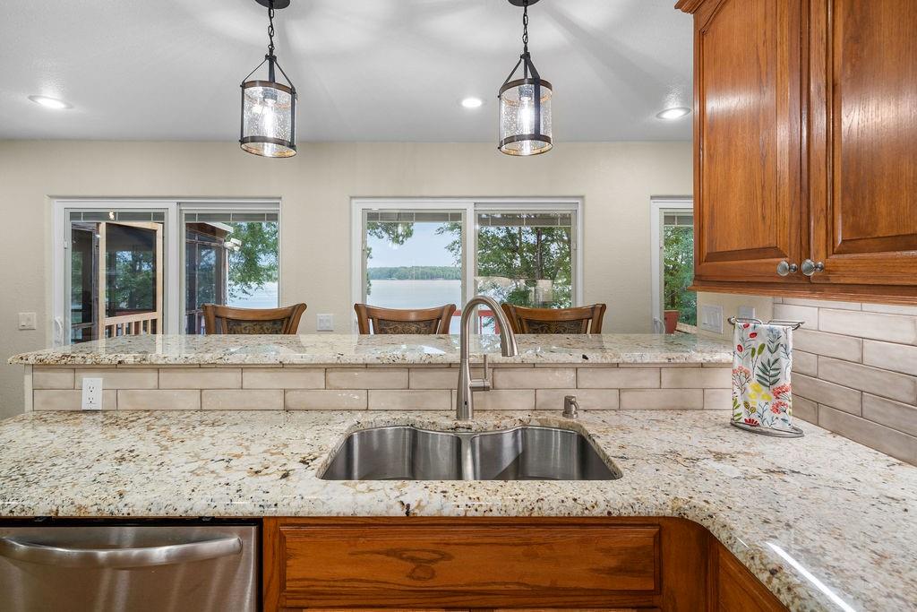 Kitchen featuring stone countertops, tile backsplash, stainless steel dual-basin sink with gooseneck faucet, stained wood cabinetry, and pendant lighting