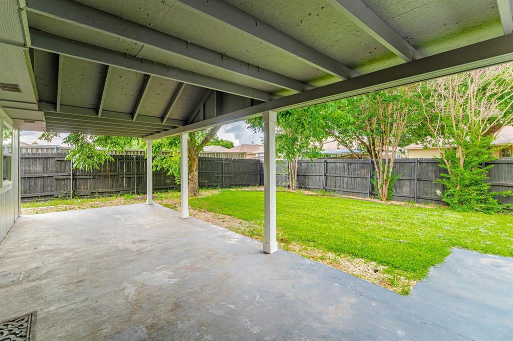 Covered concrete patio featuring exposed rafters and square support columns, opening to a fenced backyard with a lawn and mature trees