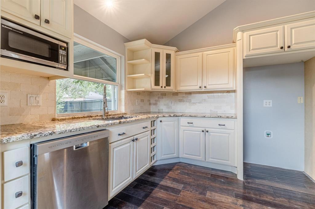 Kitchen featuring cream-toned cabinetry with glass-front display sections, stone-finish countertops, subway-style backsplash, integrated stainless steel dishwasher, and wood-finish flooring