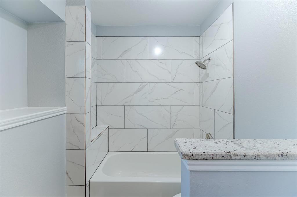 Tub and shower combination featuring floor-to-ceiling large-format tile with veining, built-in recessed shelving, and a stone-topped half wall