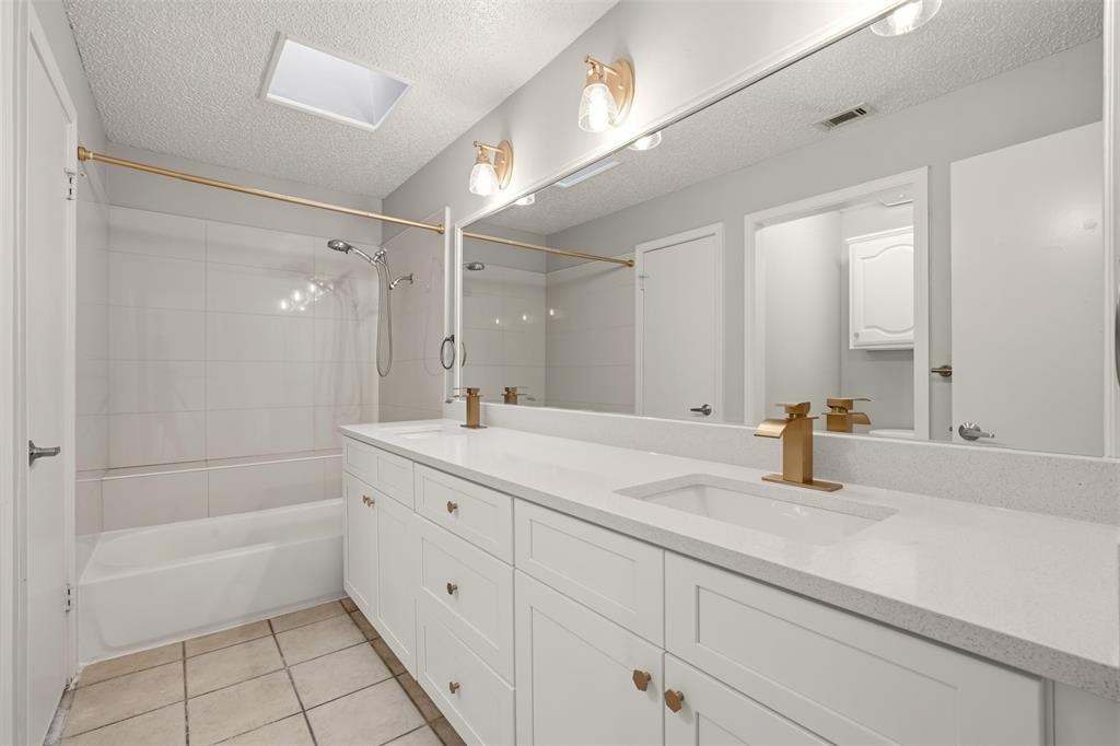Full-width vanity with white cabinetry and quartz countertop, dual integrated sinks, brass-finish hardware, and built-in skylight