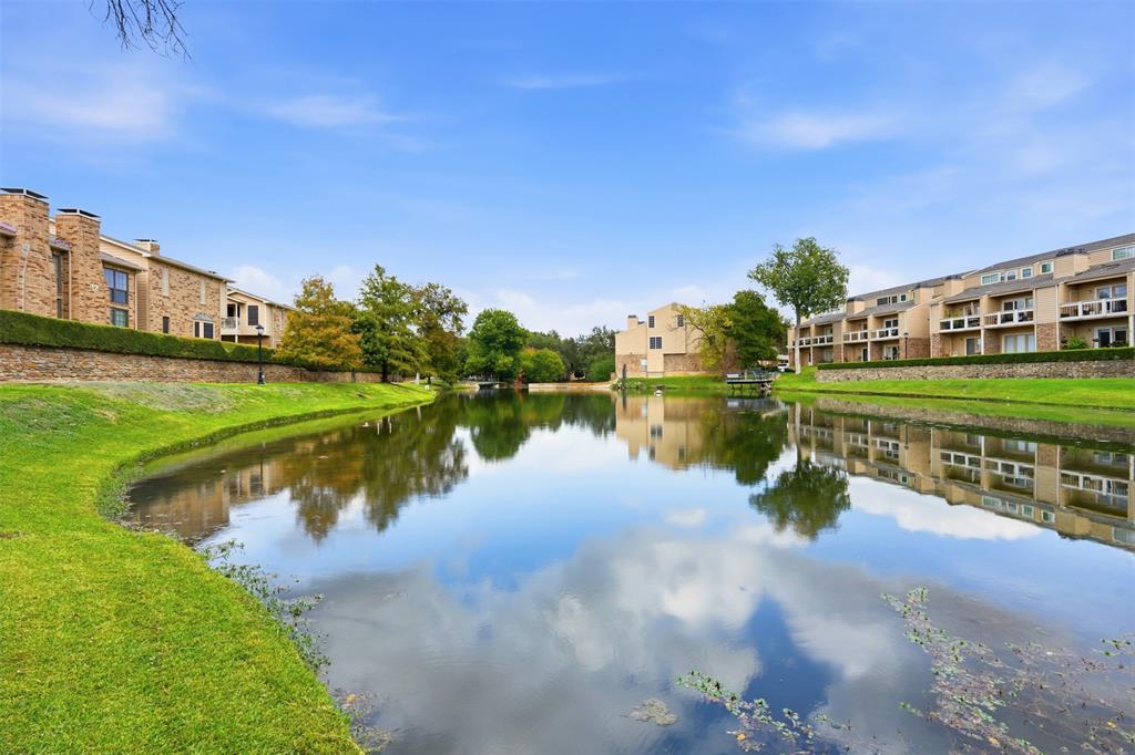 Waterfront property featuring a central pond, expansive manicured lawn, stone retaining walls, mature trees, and multi-story building exteriors with attached balconies