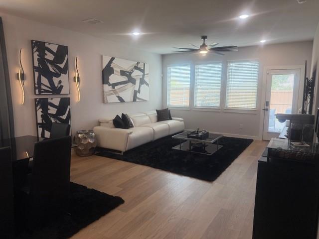 Open-concept interior featuring light-tone wood-finish flooring, recessed lighting, a modern ceiling fan, wall-mounted accent lighting, and a glass-paneled exterior door Open-concept interior featuring light-tone wood-finish flooring, recessed lighting, a modern ceiling fan, wall-mounted accent lighting, and a glass-paneled exterior door